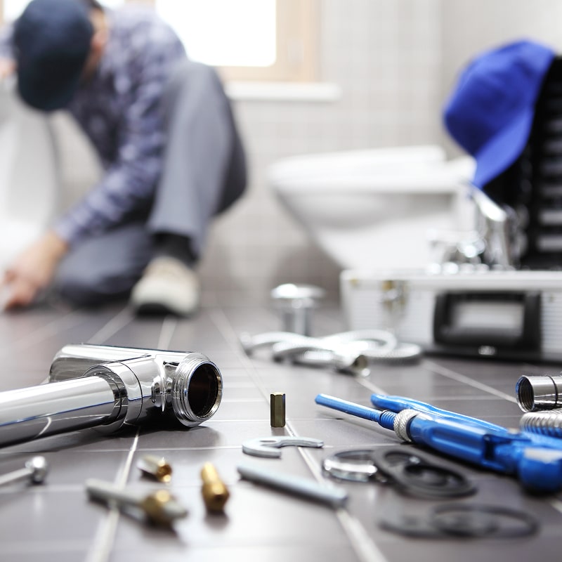 Plombier réparant des toilettes, genoux à terre, dans une salle de bain. Des outils de plomberie chromés et bleus sont éparpillés au premier plan.