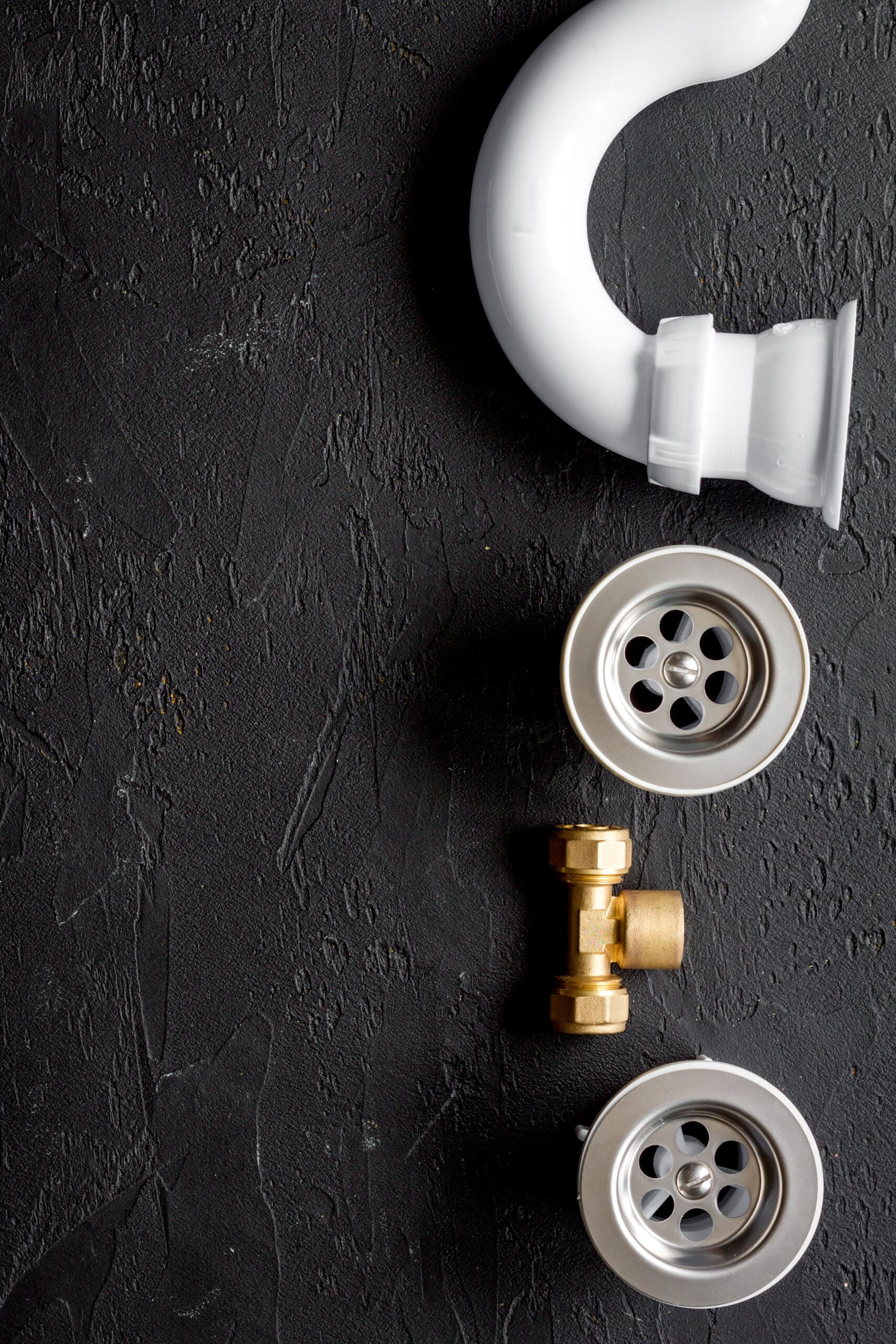 Matériel de plomberie : siphon, bondes d'évier et raccords Siphon blanc, deux bondes d'évier métalliques et raccord en laiton sur fond texturé noir. Matériel de plomberie pour installation.