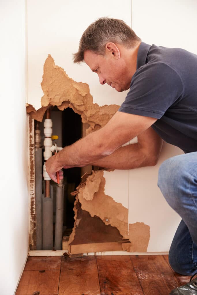 Réparation fuite d'eau : plomberie murale et dégâts Homme concentré réparant une fuite d'eau. Accroupi, il manipule des tuyaux de plomberie exposés derrière un mur cassé. Du parquet mouillé est visible.