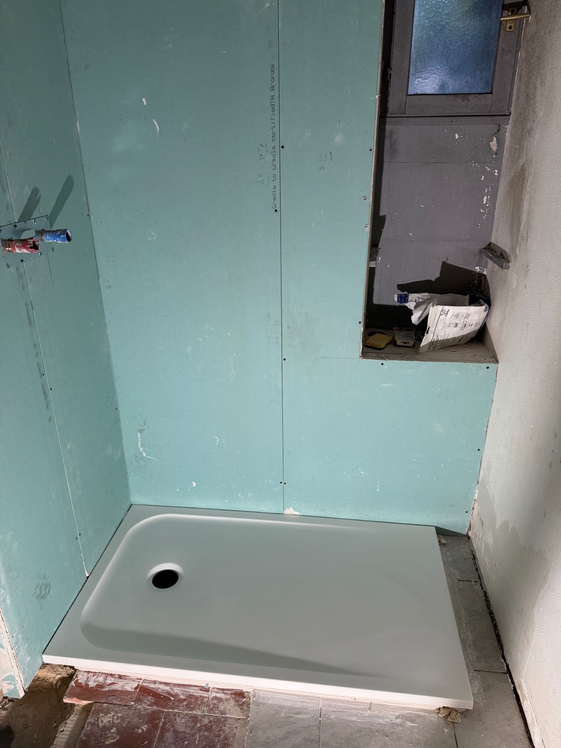 Bac à douche blanc posé contre du placo hydrofuge vert. Plomberie apparente et niche de fenêtre en cours de rénovation.