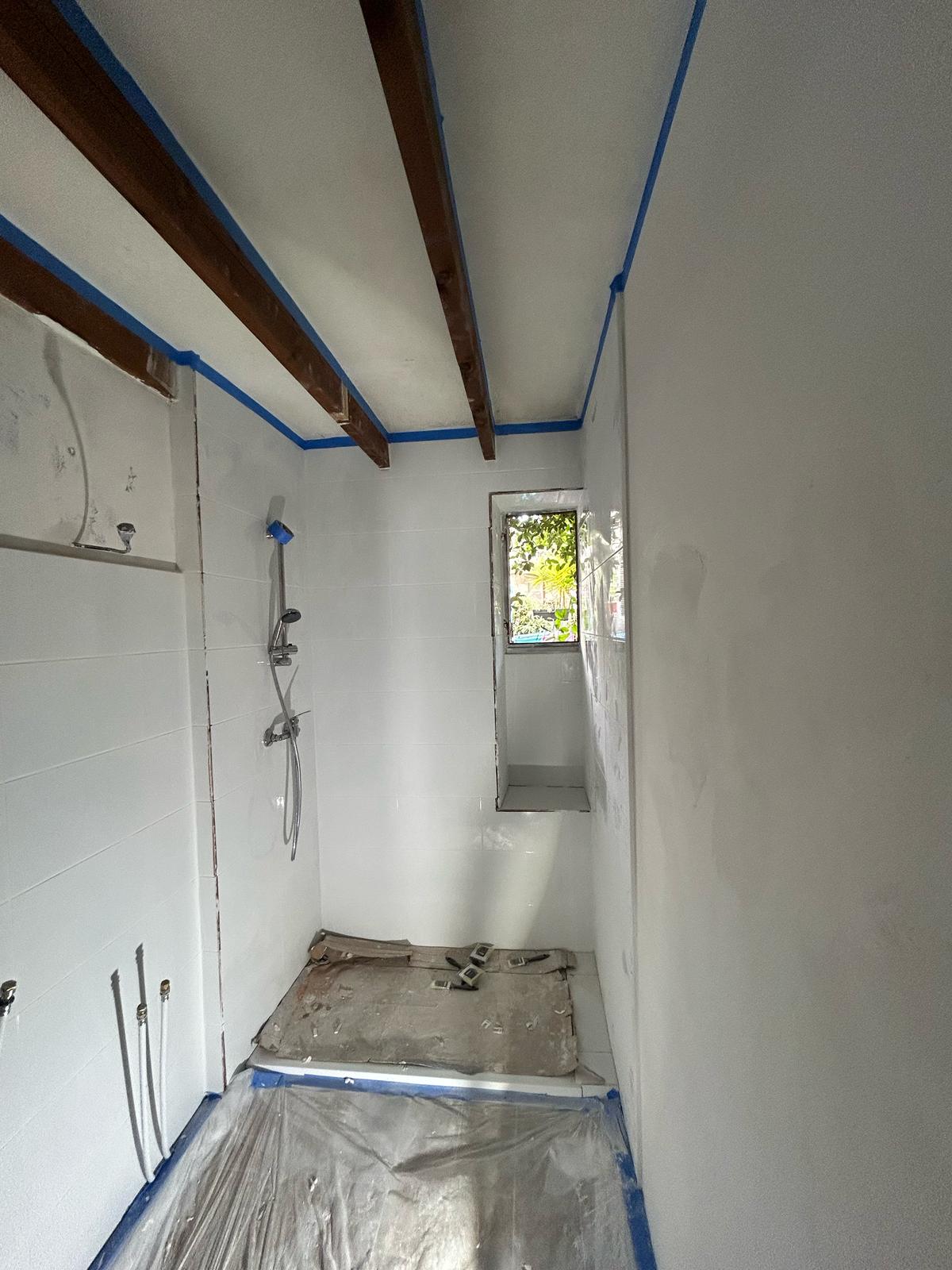 Salle de bain en chantier avec carrelage blanc, poutres en bois et douche installée.