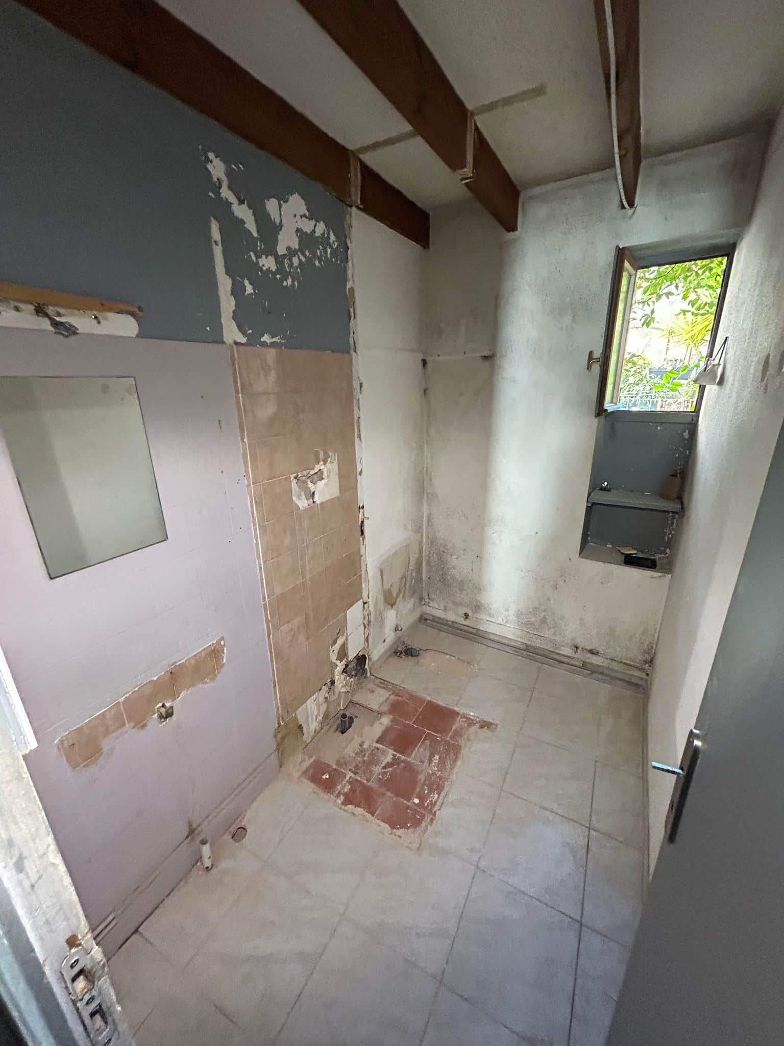 Salle de bain délabrée en rénovation, murs écaillés et poutres apparentes.