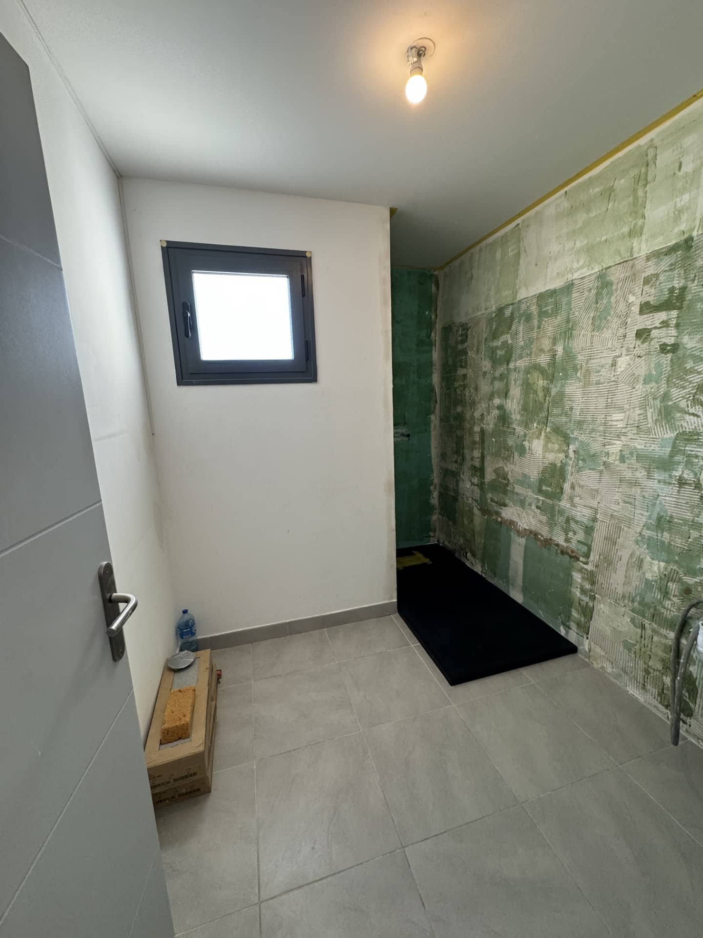 Salle de bain en rénovation : bac à douche noir, étanchéité murale verte et carrelage gris moderne au sol.