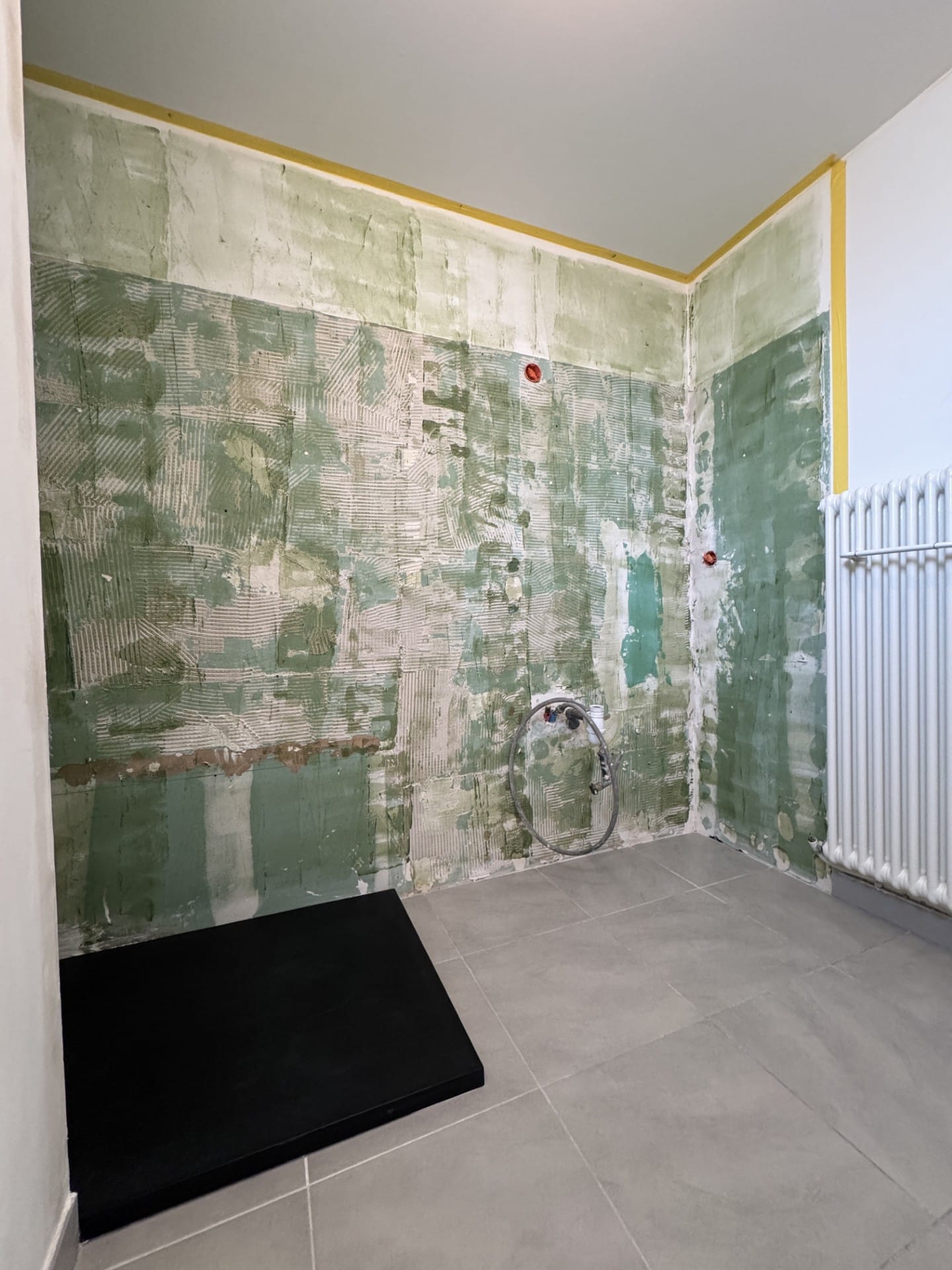 Chantier de salle de bain : murs grattés, carrelage gris et receveur de douche noir.