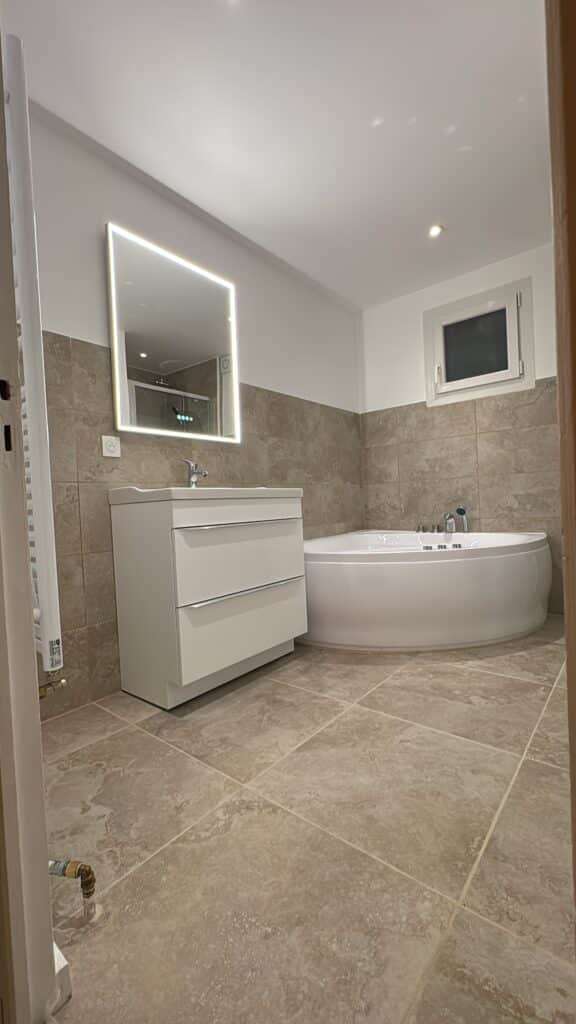 Salle de bain moderne : baignoire d'angle et miroir LED Salle de bain moderne : carrelage beige, vanité blanche, miroir rétroéclairé et grande baignoire d'angle blanche.