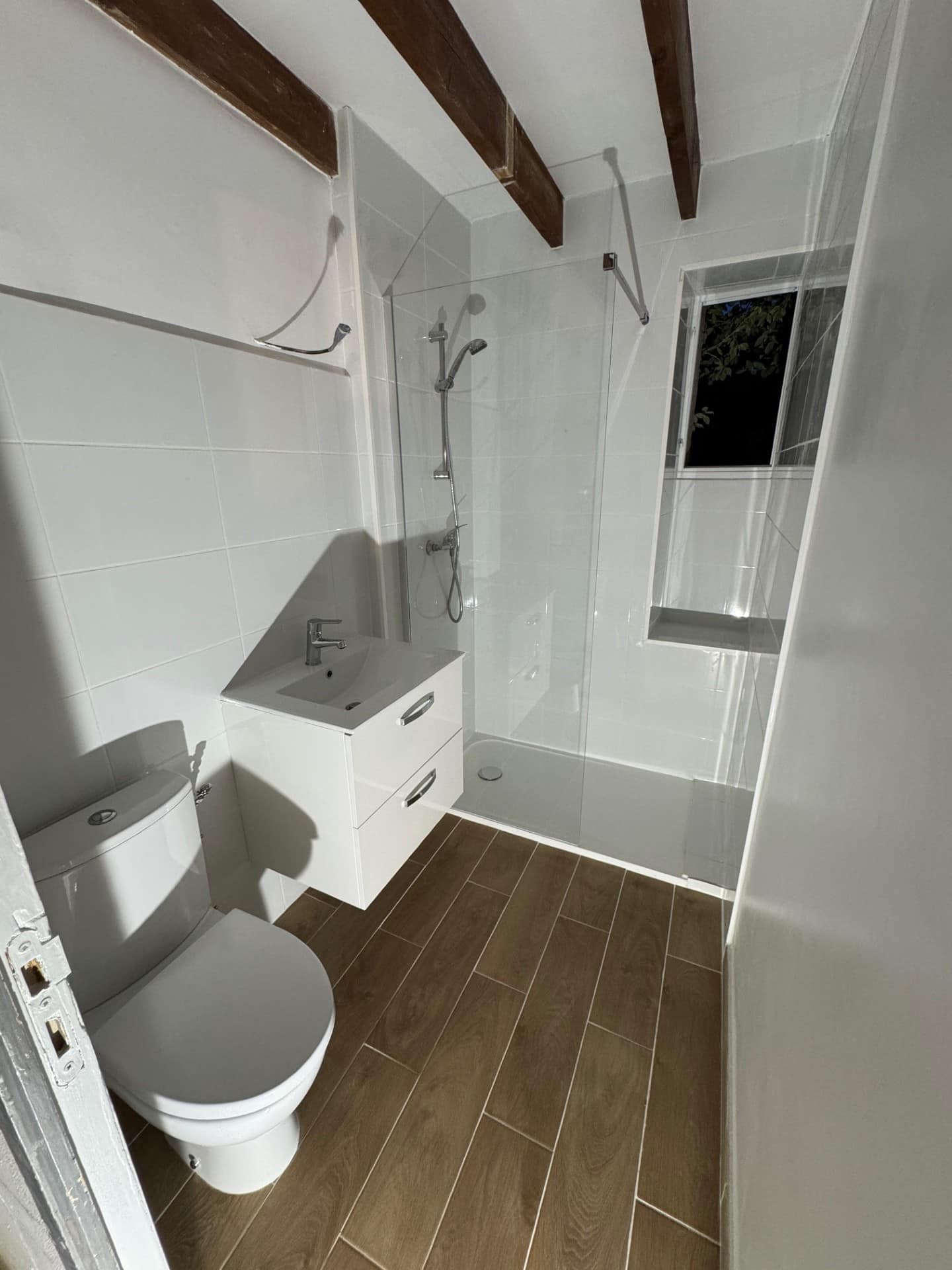 Salle de bain moderne avec douche italienne, meuble vasque blanc et sol aspect bois sous poutres apparentes.