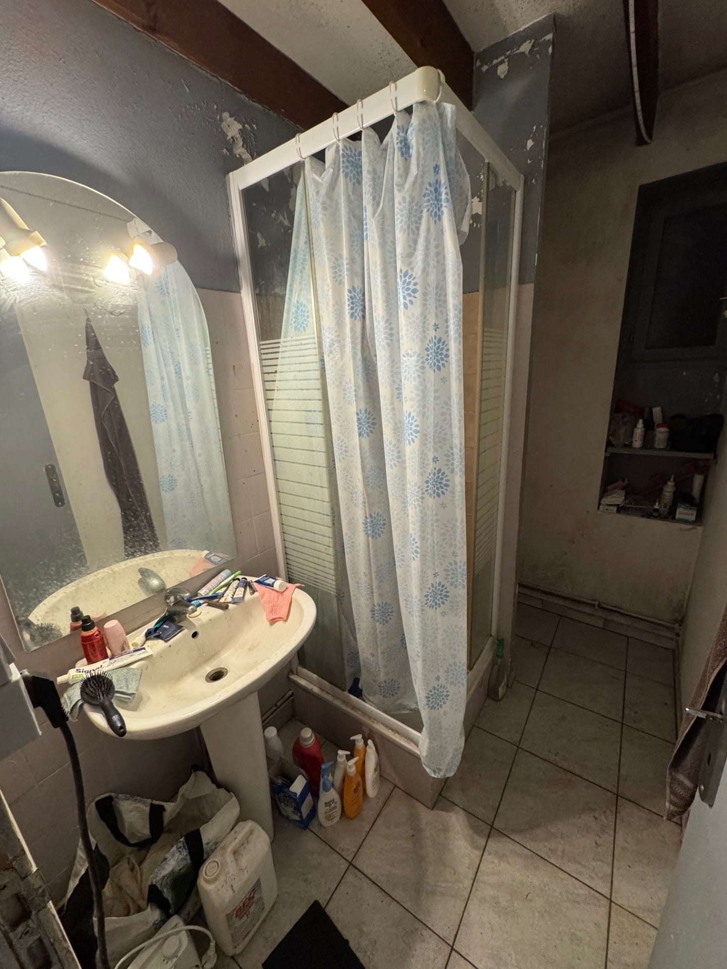 Salle de bain négligée avec lavabo encombré, douche à rideau fleuri bleu et murs à la peinture écaillée.