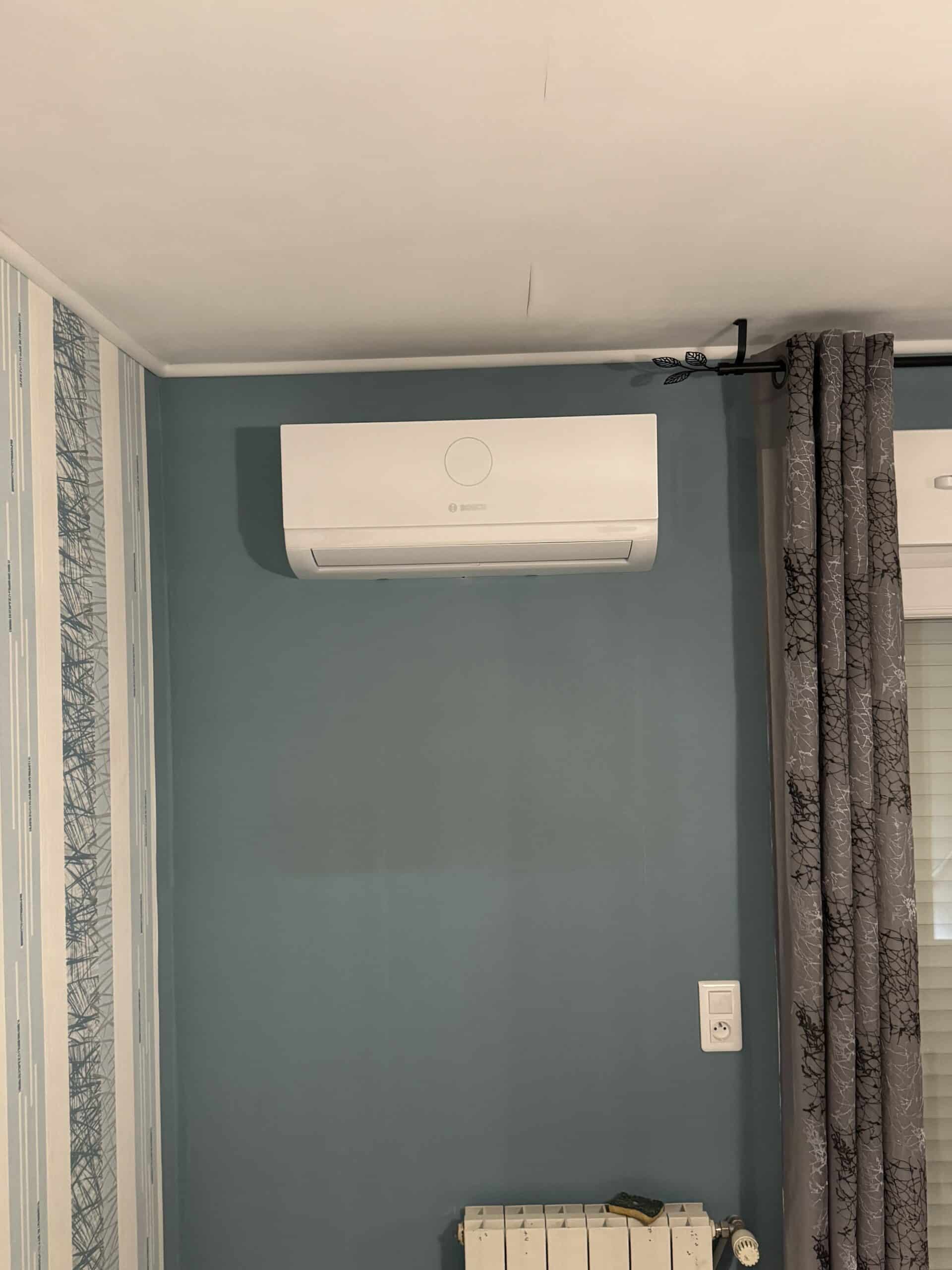 Climatiseur Bosch blanc fixé sur un mur bleu canard entre un rideau gris et un papier peint graphique.