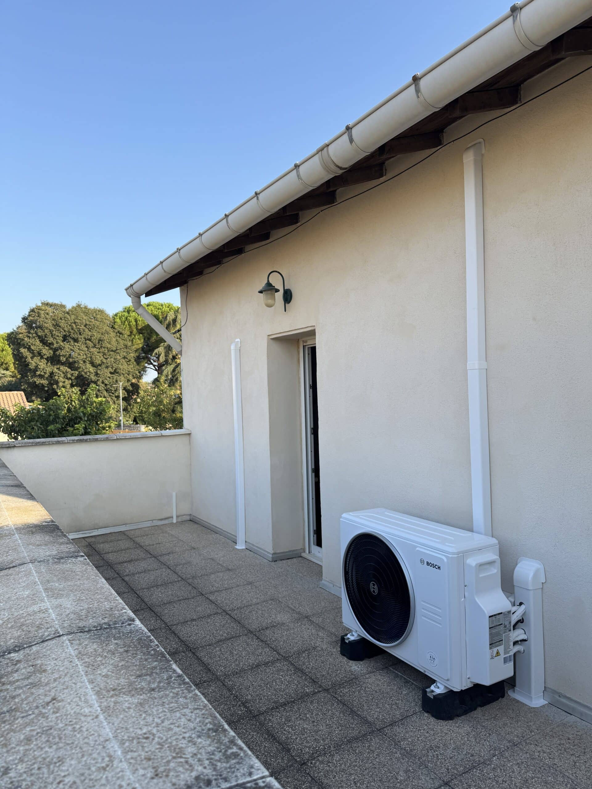 Unité extérieure Bosch blanche pour climatisation installée sur une terrasse carrelée contre un mur clair.