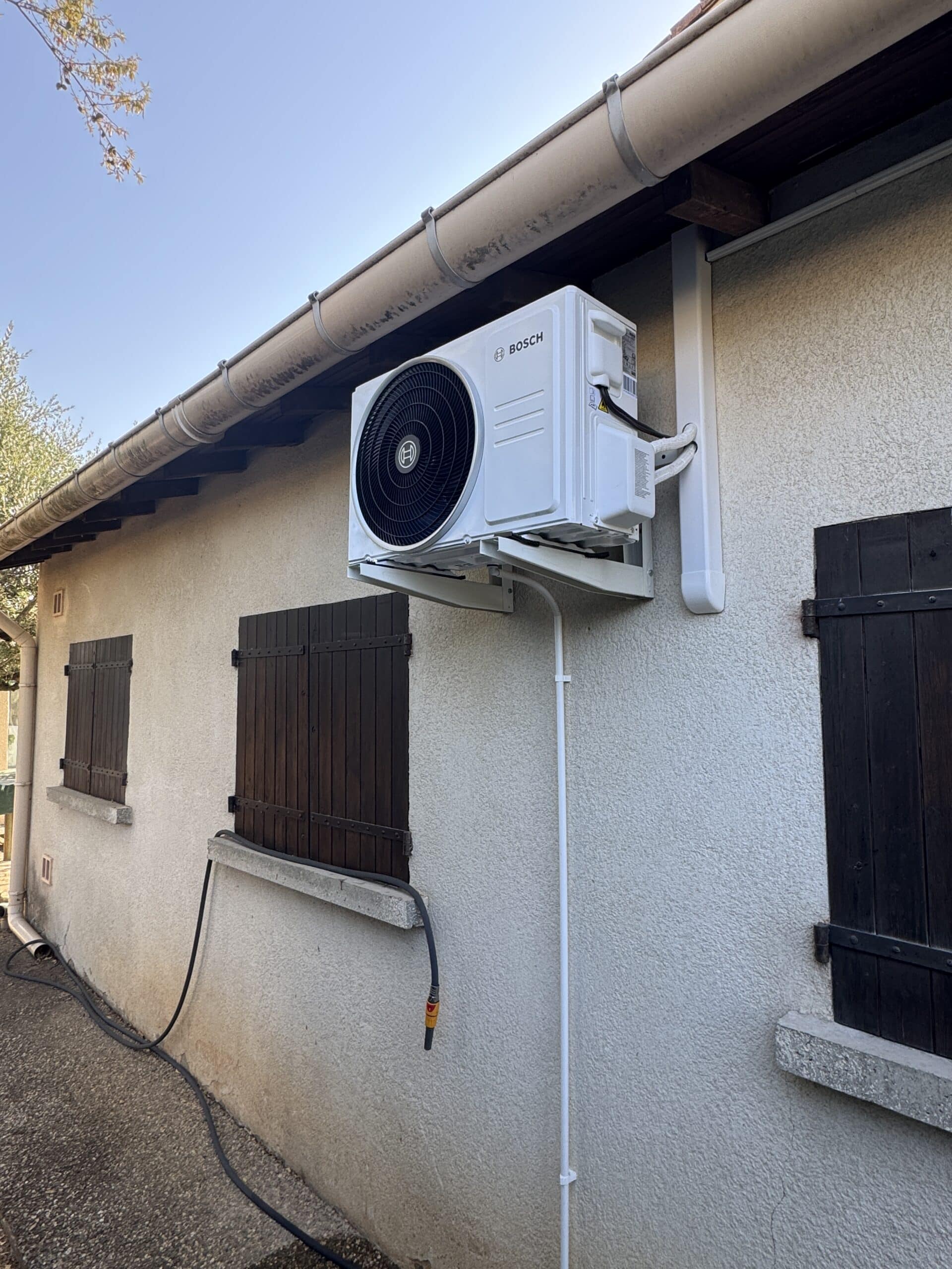 Unité extérieure de climatisation Bosch blanche installée sur une façade avec volets en bois sombres.