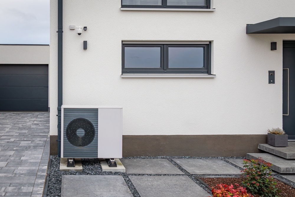 Installation d'une pompe à chaleur air-eau maison moderne Pompe à chaleur air-eau installée devant une maison moderne blanche avec fenêtres sombres et sol pavé.